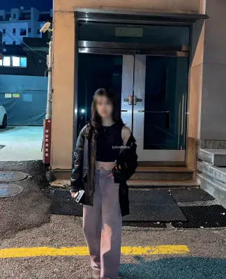 call girl in indiapuram stands outside a building at night. Theyre wearing a black leather jacket over a black top and purple pants, holding a phone. The scene is dimly lit, with a parked car and buildings visible in the background.